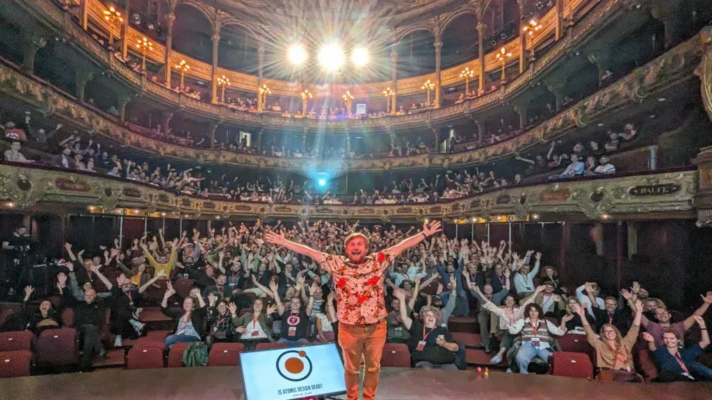 Brad Frost on stage with outstretched hands at a theater in Antwerp Belgium. All of the audience has their hands up.