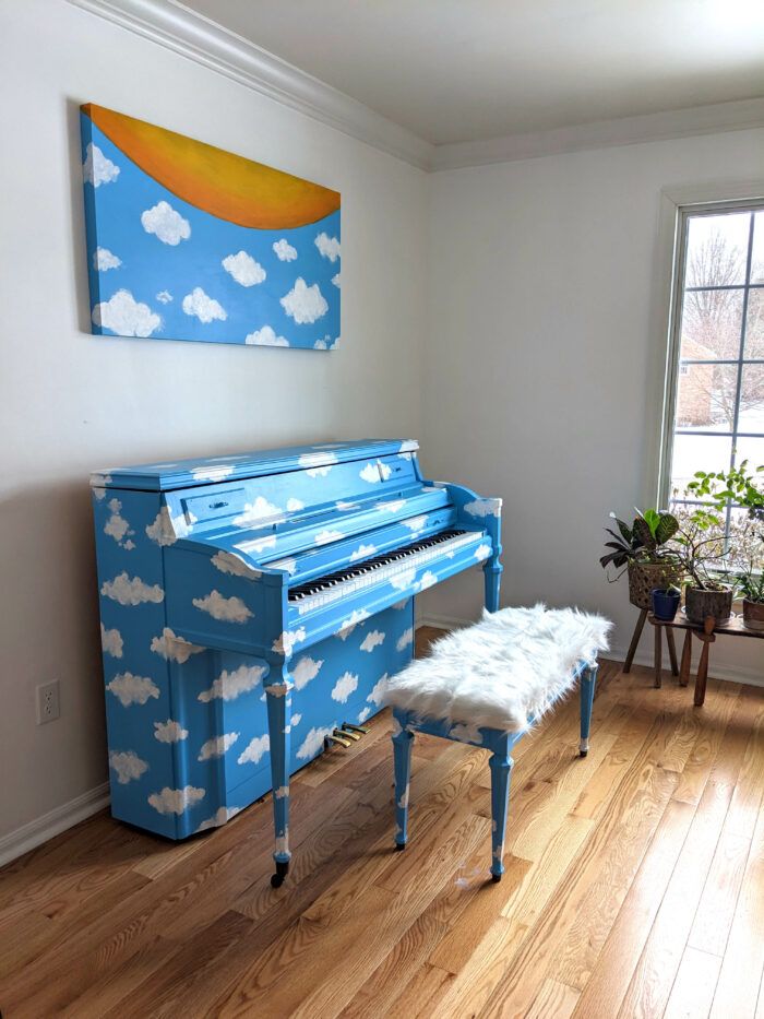 A sky blue piano with cloud pattern painted on it. A canvas with matching pattern and a semi-circle sun shape sits above the piano. A piano bench with